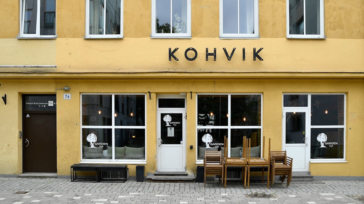 A charming and symmetric storefront of a traditional Estonian coffee shop with a vibrant yellow facade, featuring large glass windows, wooden outdoor furniture stacked up, and minimalist black signage reading "KOHVIK".📸 Photo by me IG: @margoevardson